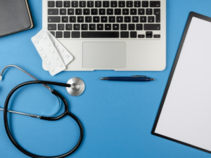 a laptop keyboard, a stethoscope, blister packs of pills, a notebook and a file on a blue background
